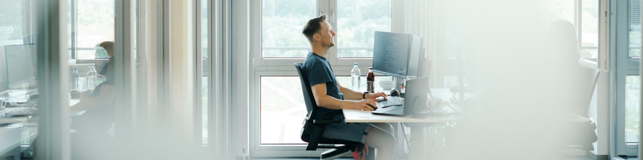 Ein Mann sitzt an einem Schreibtisch und arbeitet an einem Computer in einem modernen, hellen Büro mit großen Fenstern. Er lacht. Eine weitere Person ist in einem separaten Büroraum im Hintergrund zu sehen.