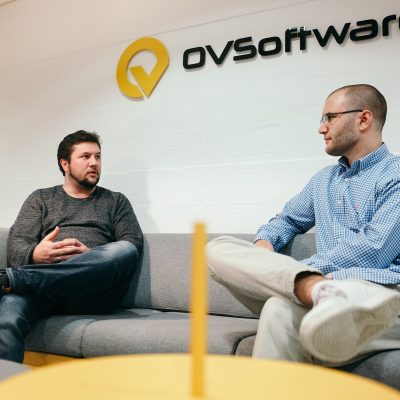 Zwei Softwareentwickler sitzen auf einer grauen Couch und führen ein Gespräch in einem modernen Bürogebäude. Hinter ihnen ist das Logo von OVSoftware zu sehen. Ein Mann trägt einen Pullover und Jeans, der andere eine Brille, ein blaues Hemd und weiße Turnschuhe.